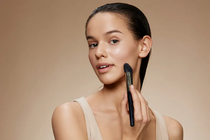 Young woman applying makeup with a brush, showcasing natural beauty with a neutral background, perfect for beauty and skincare products