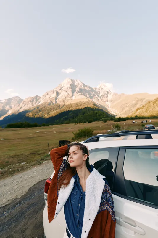 Woman by car parked in alpine mountain landscape during travel roadtrip, portrait of relaxed traveler leaning on vehicle, nature adventure with scenic horizon and sunlight.