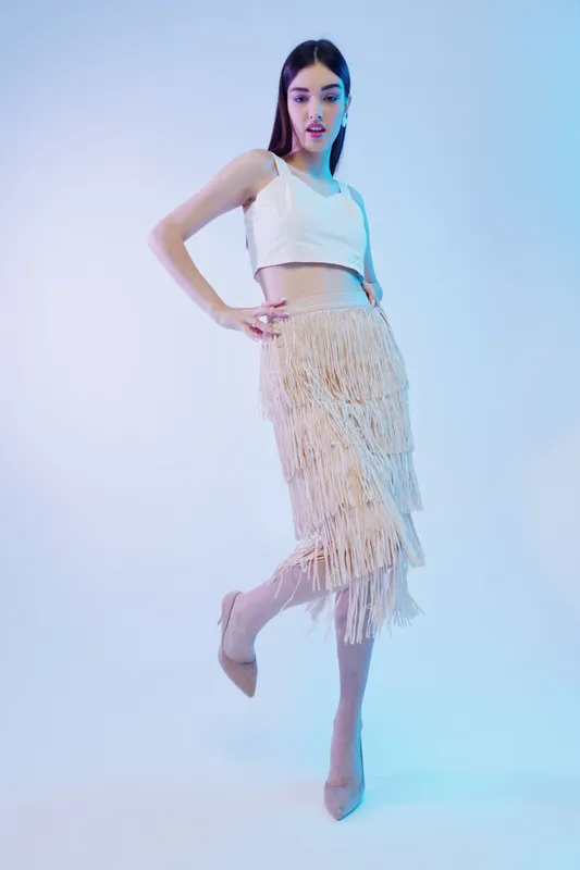 Young woman in fashionable white crop top and fringed skirt poses gracefully on one leg. Studio shot with soft blue lighting. Concept of woman beauty fashion style.