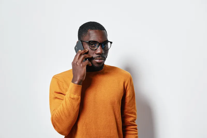 African American man with dark skin and glasses talking on smartphone wearing orange sweater against white background. Casual communication, technology and portrait with confident expression.