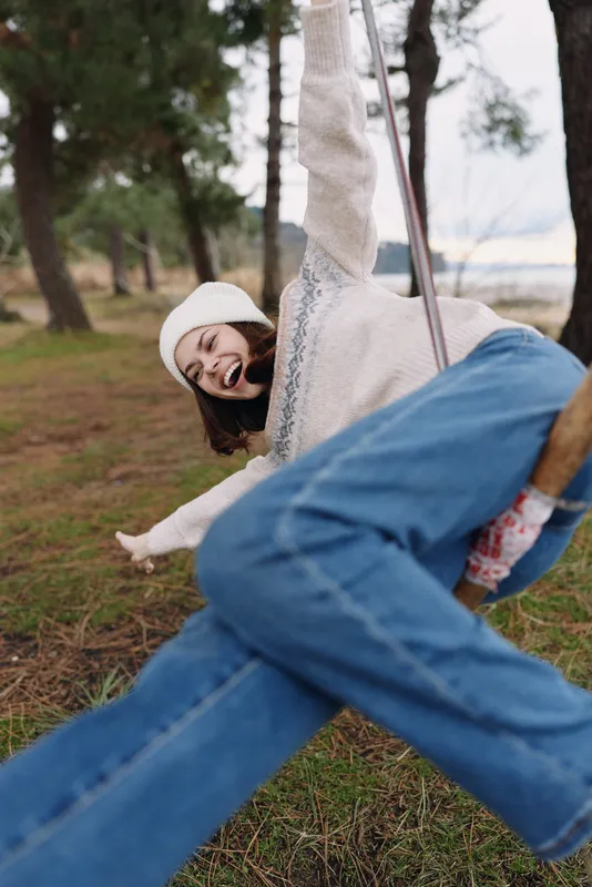 Outdoor playful girl on a swing in a park, casual denim and sweater, laughing as she enjoys nature and leisure in a sunny autumn afternoon.