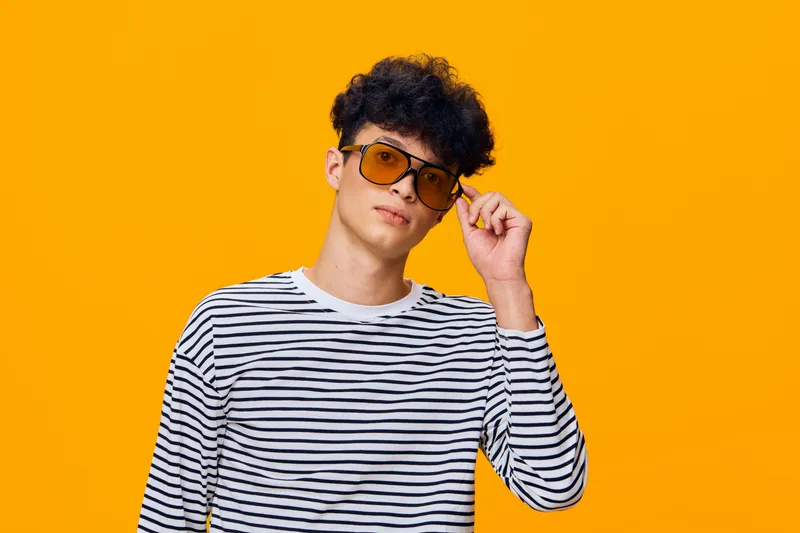 young man wearing striped shirt and tinted sunglasses posing confidently against vibrant orange background, casual style with trendy hairstyle and thoughtful expression