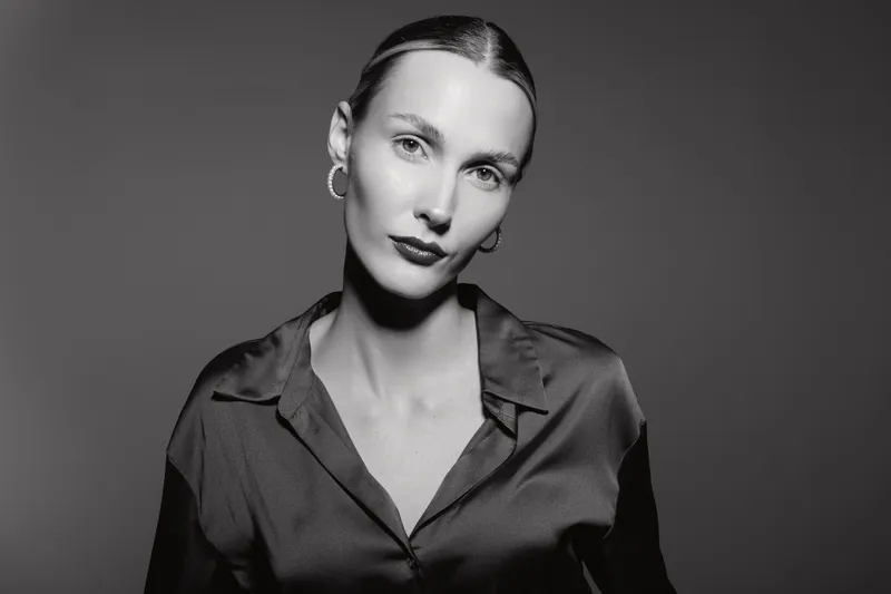 Portrait of a young caucasian woman with pale skin wearing a silk blouse and hoop earrings. Studio lighting highlights her calm expression on a dark background.