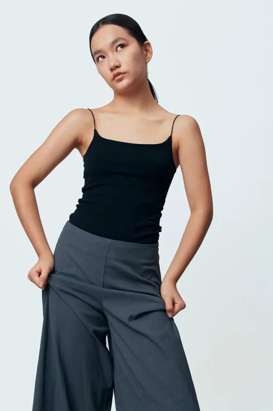 Young woman standing with hands in pockets wearing black sleeveless top and gray wide leg pants against white background. Casual fashion, confident pose, minimal outfit, studio portrait.