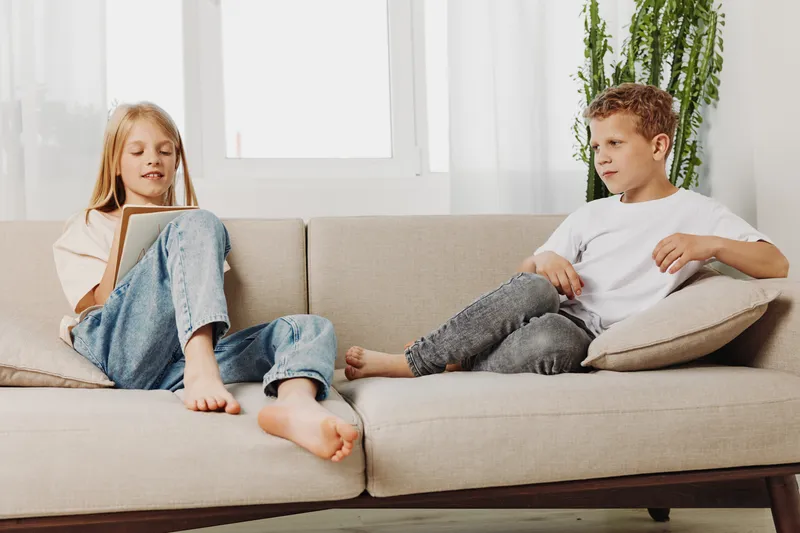 Two children relax on beige sofa in bright living room, girl reading book and boy holding glasses. Concept for childhood leisure, home comfort, and sibling interaction.