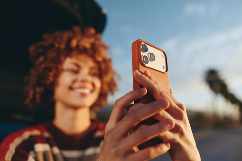 Woman lifestyle outdoors holding smartphone for selfie, curly hair smiling in soft daylight, shallow depth of field, casual red jacket, warm natural colors, relaxed street background