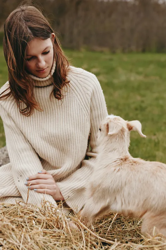 Woman in a sweater interacts with a goat kid outdoors in a field, countryside scene with nature tones, gentle moment, calm expression, rustic charm, authentic lifestyle