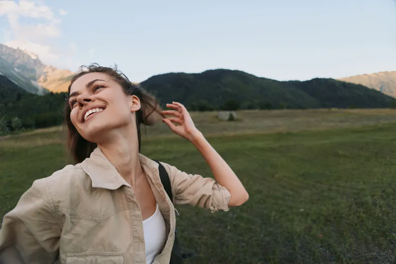 Young woman outdoors in a wide grassy field with distant mountains, smiling and flipping her hair, wearing a casual beige jacket and white top, sunlit moment