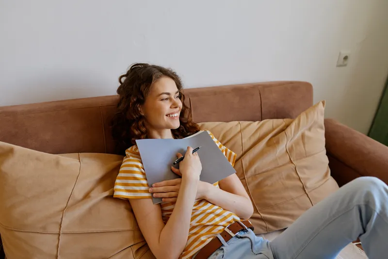 A young woman with curly hair relaxes on a sofa at home, smiling as she writes in a notebook, capturing a casual, comfortable lifestyle moment in a bright living room.