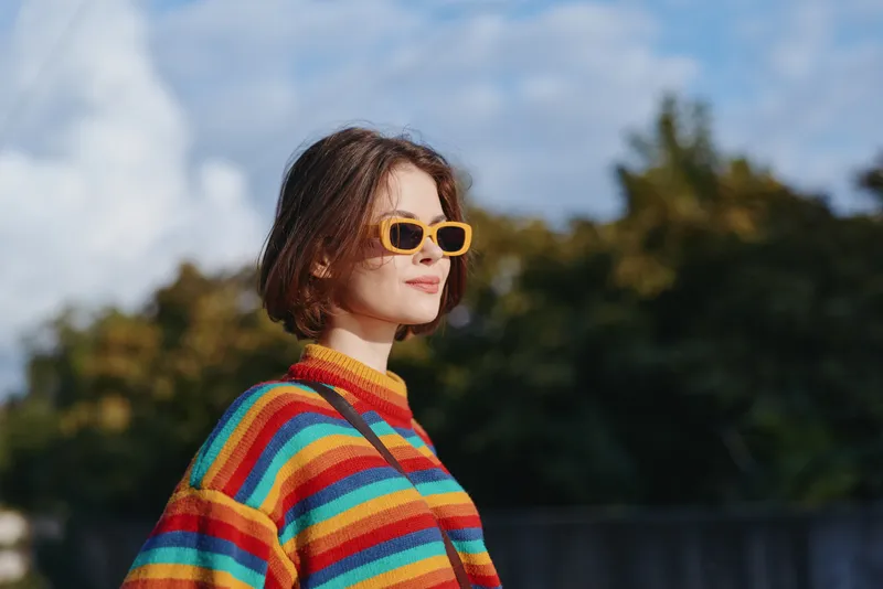 Woman in sunglasses and colorful striped sweater standing outdoors in sunny portrait, short hair and confident pose, casual fashion style and youthful street look.