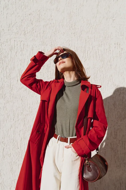 One stylish woman wears a red trench over an olive green top and white trousers, standing against a pale wall with a sunlit, relaxed pose.