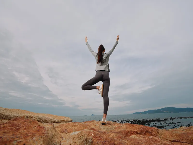 Female model practicing yoga on rocky beach, showcasing balance and tranquility against a serene ocean backdrop, promoting mindfulness and healthy lifestyle