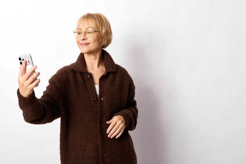 Woman with smartphone, elderly person with glasses, smiling during studio portrait indoors, soft light, neutral background, relaxed pose, hands visible