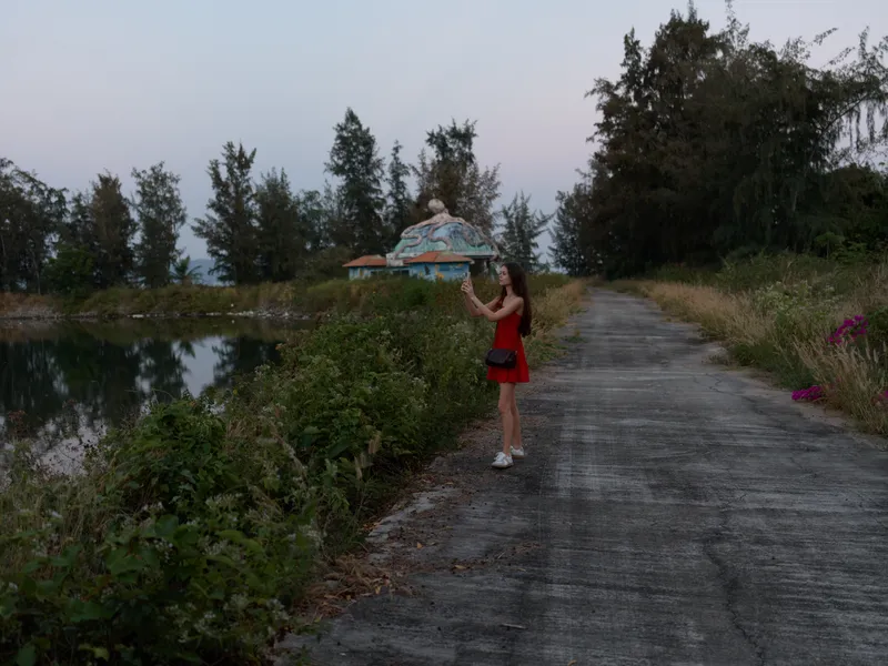 Young woman in red dress taking a photo with smartphone near a calm lake at dusk, standing on a quiet rural road surrounded by trees and greenery.