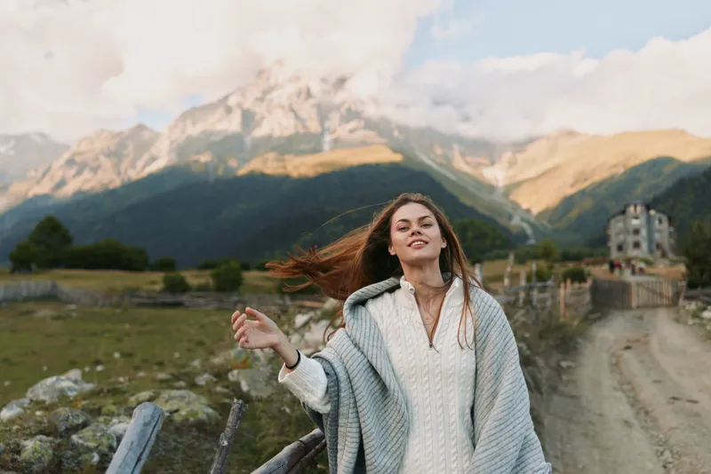 Young woman in a cozy sweater poses outdoors in a rugged mountainscape, wind catching her hair as she enjoys a carefree moment surrounded by nature.
