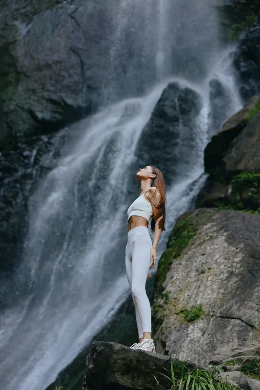 young woman standing near waterfall in white activewear, enjoying nature outdoors with serene expression and fitness vibe in lush green environment