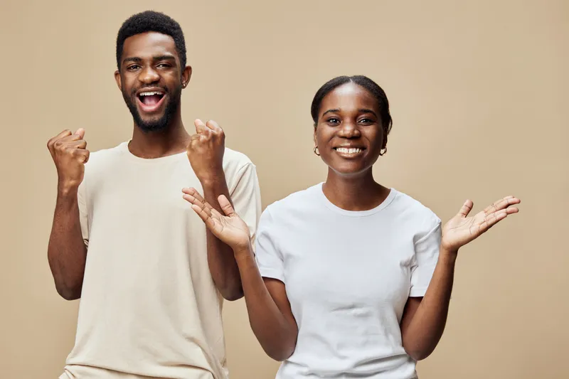 Happy young couple celebrating success with joyful expressions on a neutral beige background, embodying positivity and enthusiasm in casual clothing