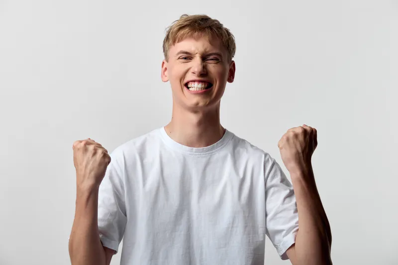 Joyful, young man in a white tee raises fists in a victory gesture, expressing celebration, happiness, and triumph during a studio shoot with an isolated background