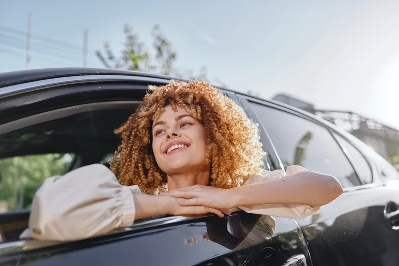 Smiling woman with curly hair enjoys sunlight leaning out car window, wearing beige blouse, conveying happiness and freedom outdoors on a sunny day.