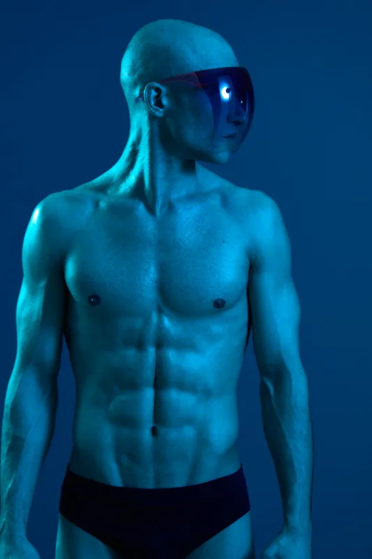 Athletic man posing confidently in a blue light, showcasing strong muscles and unique sunglasses, creating a striking visual impact.