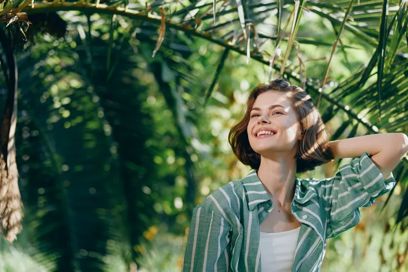 Young woman smiling in sunlit tropical garden, wearing a striped shirt, relaxed and carefree, capturing a bright portrait outdoors.