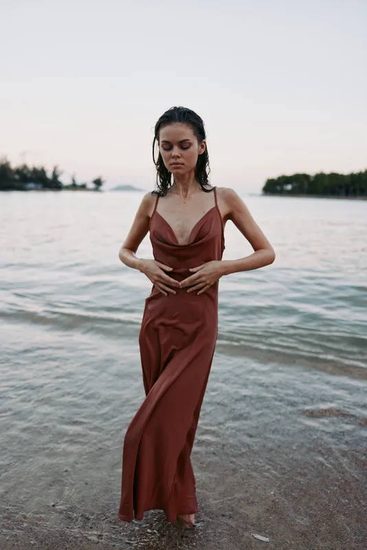 Woman in a rust red slip dress stands in shallow seaside water, hands at her waist, calm gaze forward, subtle breeze creating gentle movement.