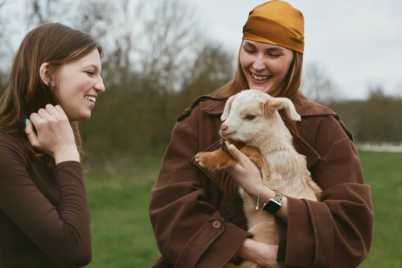 Cheerful woman outdoors holding a kid goat, sharing a friendly moment in a sunlit field, rustic jackets, natural colors, warm breeze, candid laughter, countryside mood, fieldside joy