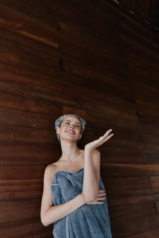 smiling woman wrapped in towel with hair towel wrap against wooden wall, natural skincare, morning routine, spa day relaxation, fresh and joyful mood