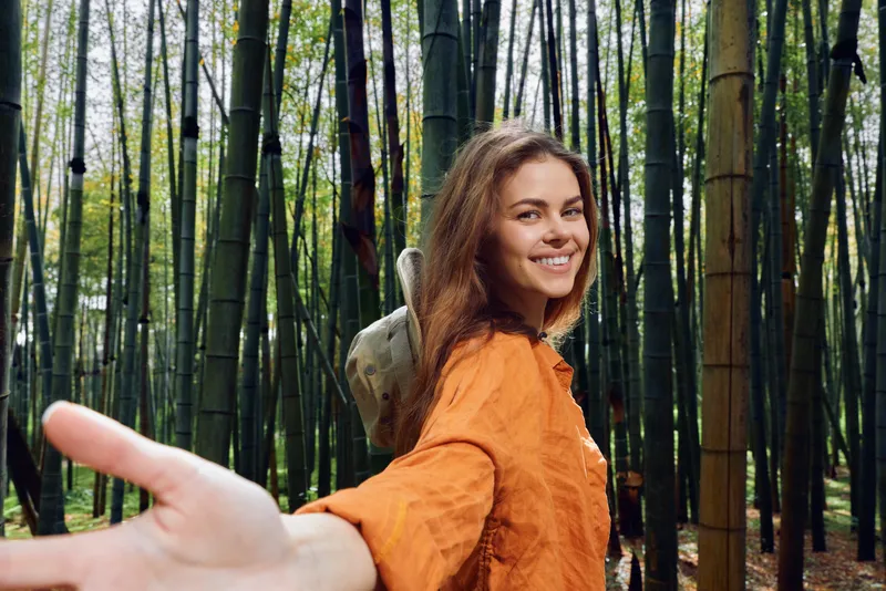 A smiling woman traveler in a bamboo forest reaches out holding a hand, portrait and nature scene with orange dress, joyful expression, candid outdoor adventure and green foliage moment.