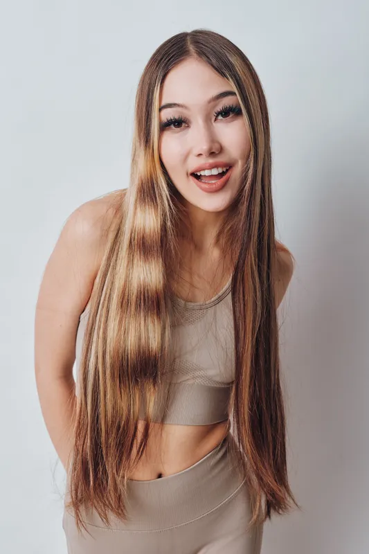 Young woman with long straight hair wearing casual outfit smiling and posing against light background in studio. Positive girl with natural makeup and relaxed posture.