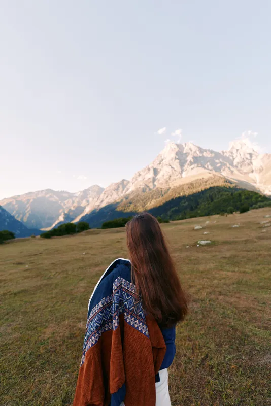 Woman back view standing in meadow, looking at snow-capped mountains and wide landscape. Nature travel scene with long hair, coat draped over shoulder and hiking mood.