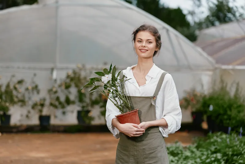 Young female gardener holding a potted plant in a greenhouse, showcasing her love for nature and sustainability in a serene outdoor setting