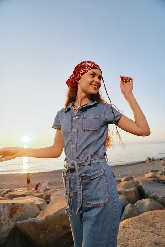 smiling woman wearing denim dress and red bandana standing on rocky beach at sunset with ocean background