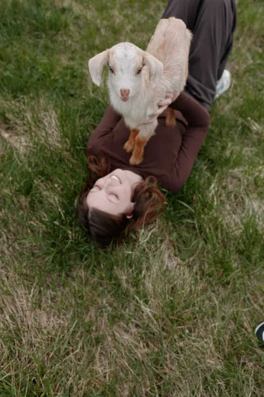 Goat kid rests on a smiling woman in outdoors grass, a wholesome lifestyle moment with playful interaction, sunlit scene, gentle breeze, natural colors, serene countryside backdrop