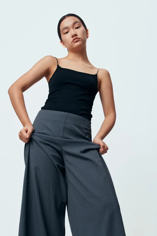 Young woman in black tank top and wide gray pants poses confidently against white background in fashion portrait studio. Minimalist style with strong attitude and casual outfit.