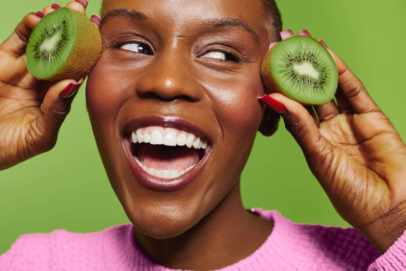 Happy dark skinned African woman with short hair holds two kiwi halves near face smiling broadly on green background. Concept of healthy lifestyle, fresh fruit, and natural beauty.