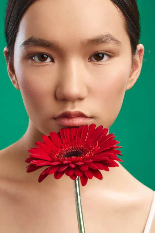 Asian woman is captured in a closeup portrait holding a vibrant red flower, set against a bold green backdrop. The expression exudes calm poise and gentle charm, creating a serene, modern aesthetic
