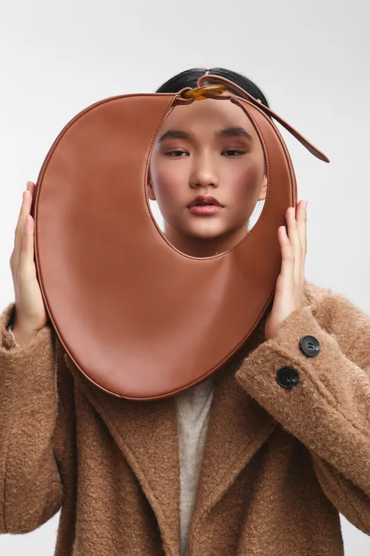 Asian woman wearing a bold circular leather neckpiece with a textured coat, creating a striking fashion statement in a studio setting, posed confidently with soft lighting and calm gaze.