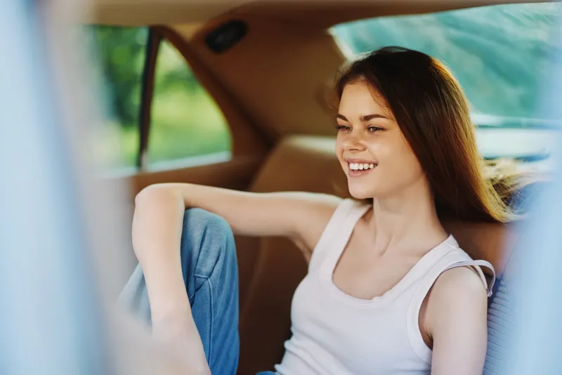 Smiling young woman sitting in a car, wearing a casual white tank top and jeans, exuding happiness in a natural outdoor background.