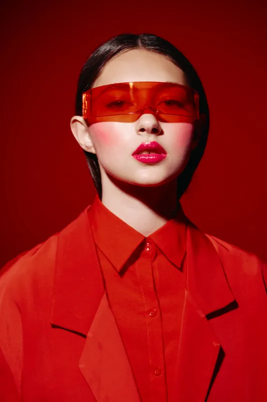 Young woman in vibrant red jacket and shirt wears futuristic orange sunglasses on red background. Bold makeup and hairstyle enhance fashion and beauty mood.