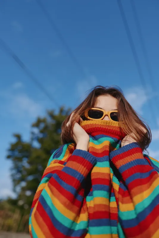 young woman in a colorful striped sweater and sunglasses outdoors, playful pose pulling collar over mouth under blue sky. Teen model with long brown hair and cozy oversized knit vibe.