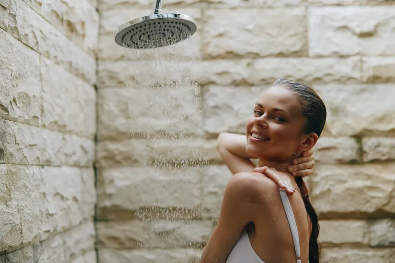 smiling woman enjoying outdoor shower with wet hair against stone wall, natural light, fresh and relaxed mood, summer wellness moment, healthy lifestyle