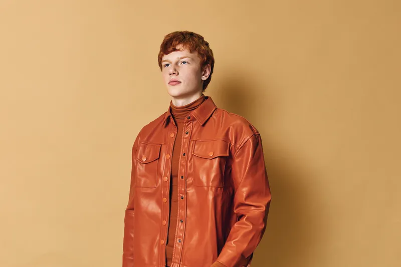 Young man with red hair wearing orange leather jacket and turtleneck stands against beige background looking forward with calm serious expression.
