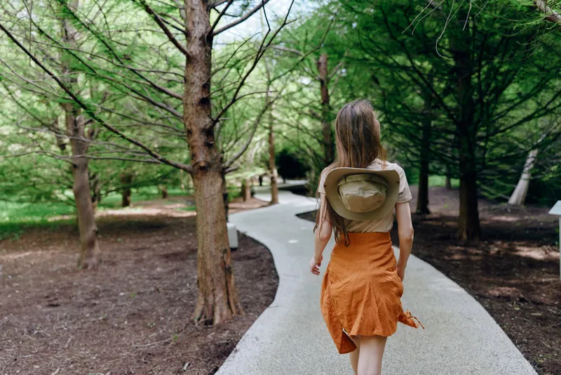 woman walking path through forest trees, backview of female traveler in orange skirt with hat on back, nature trail in park, summer stroll and peaceful outdoor scene