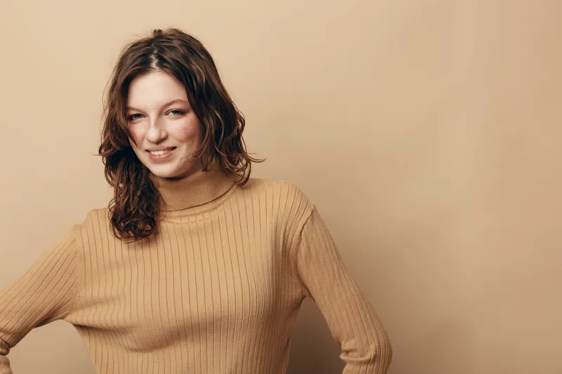 Smiling young woman with wavy brown hair wearing a beige knitted sweater stands against a neutral background. Concept of casual style, natural beauty, and relaxed mood.