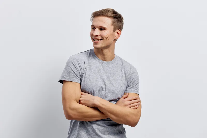 Smiling young man with casual gray t-shirt crossing arms, looking away with confident and cheerful expression isolated on plain white background. People lifestyle concept, positive mood.