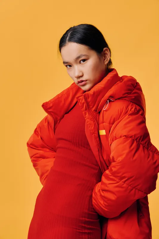 Young woman posing in red winter jacket and dress looking at camera with confident expression on yellow background. Fashion, style, portrait and casual wear for cold season clothing.