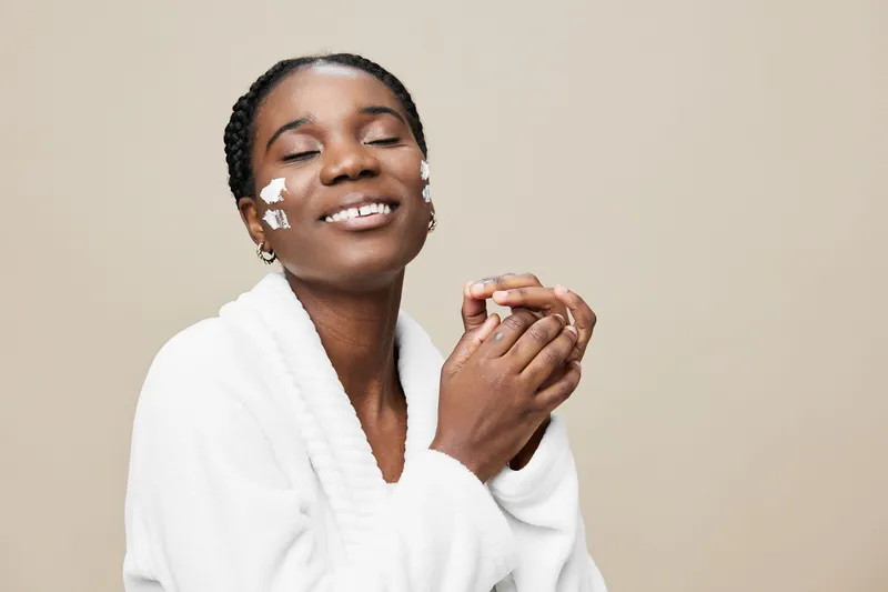 Young Black woman with a radiant smile, wearing a cozy white robe, enjoying self care with skincare products against a neutral backdrop, evoking feelings of wellness and beauty