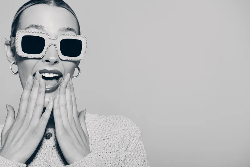 Surprised young Caucasian woman with light skin wearing large sunglasses and a knitted sweater covers her mouth with hands on a plain background.