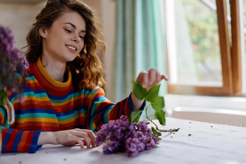 A woman in a colorful striped sweater arranges lilac flowers on a sunlit kitchen table, capturing a calm home moment and creative hobby in a cozy domestic setting.